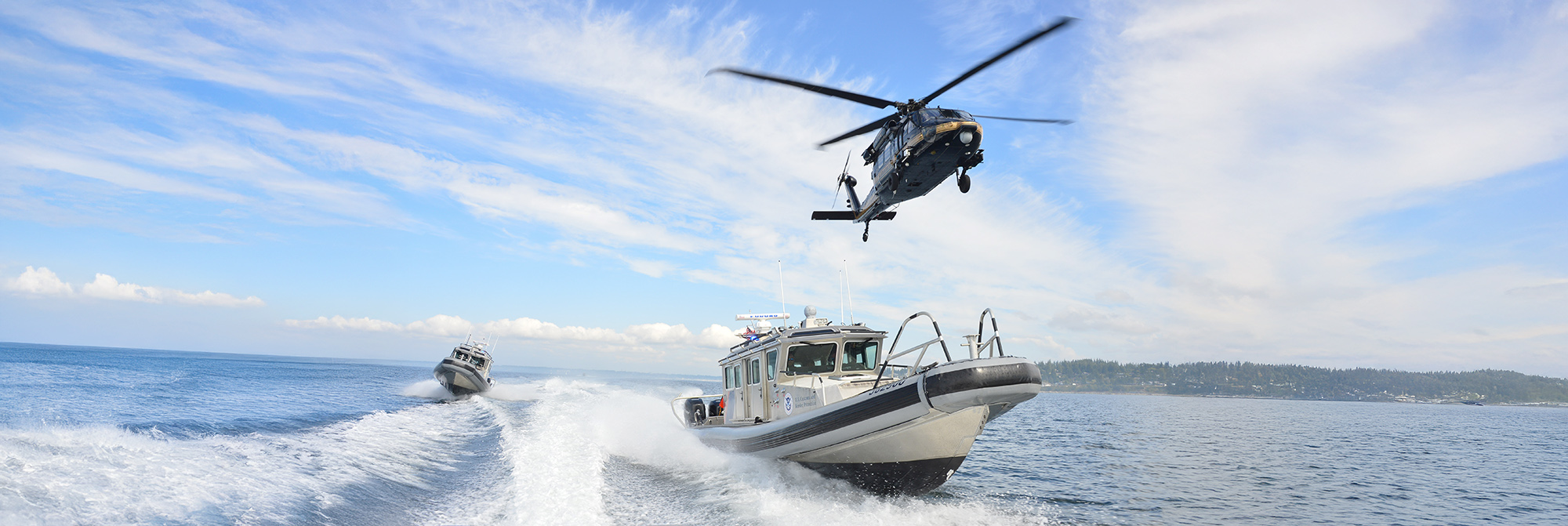 An AMO UH-60 Black Hawk flies over two All-Weather Interceptor vessels.