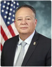 Pete Flores, Senior Official Performing the Duties of the Commissioner, seated in front of a blue background with a flag to the left.